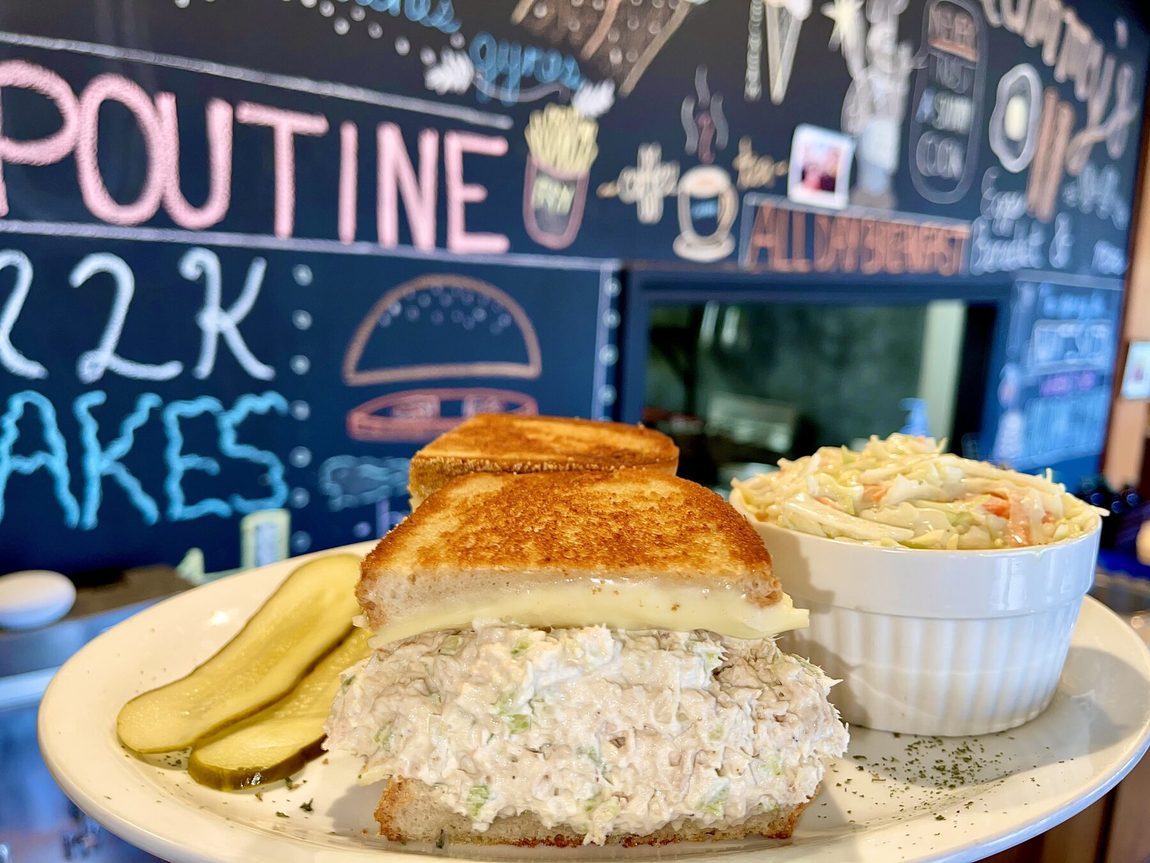 A tuna salad melt with coleslaw and pickles on the counter, chalkboard menu behind